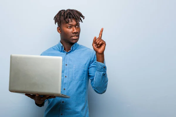 Young rasta black man holding a laptop smiling cheerfully pointing with forefinger away.