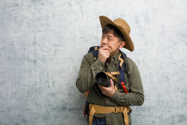 Young chinese explorer man holding a camera doubting and confused