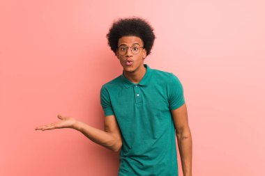 Young african american man over a pink wall holding something on palm hand clipart