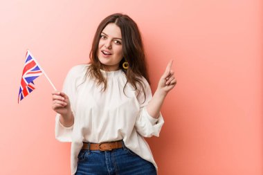Genç ve cüsseli bir kadın birleşik krallık bayrağını tutuyor ve mutlu bir şekilde işaret parmağıyla işaret ediyor..