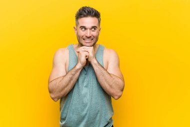 Sarı arka planda genç bir fitness adamı ellerini çenesinin altında tutuyor, mutlu bir şekilde yana bakıyor..
