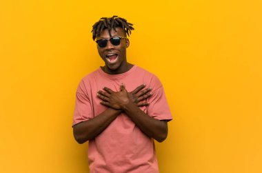 Güneş gözlüğü takan genç siyahi bir adam gülüyor ellerini kalbe koyuyor, mutluluk kavramını koruyor..