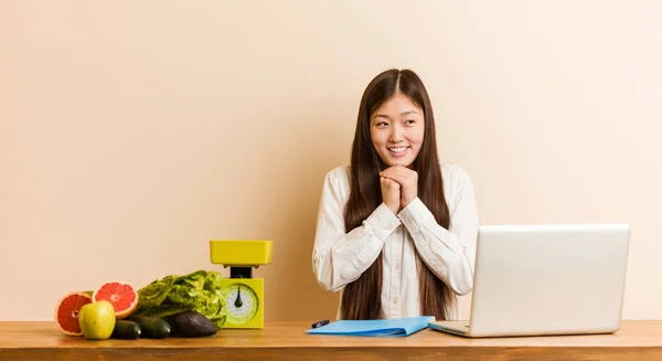 Young nutritionist chinese woman working with her laptop keeps hands under chin, is looking happily aside.