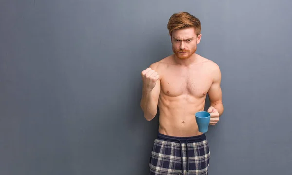 Young shirtless redhead man showing fist to front, angry expression. He is holding a coffee mug.
