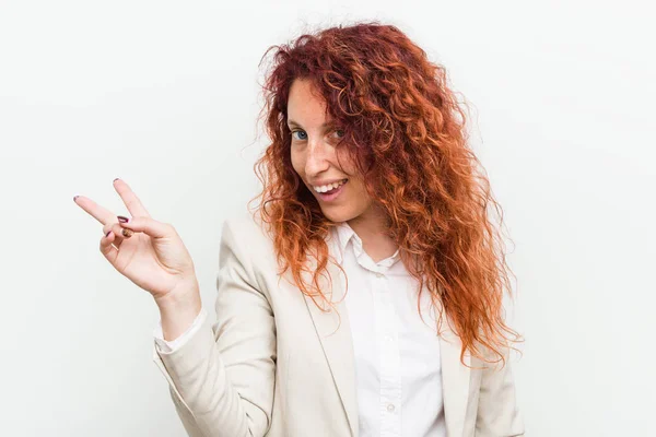 Young natural redhead business woman isolated against white background joyful and carefree showing a peace symbol with fingers.