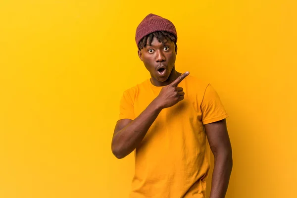 Young black man wearing rastas over yellow background pointing to the side