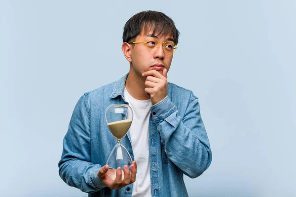 Young chinese man holding a sand timer doubting and confused