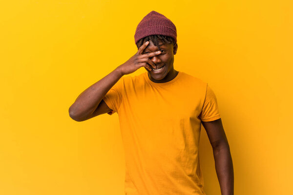 Young black man wearing rastas over yellow background blink at the camera through fingers, embarrassed covering face.