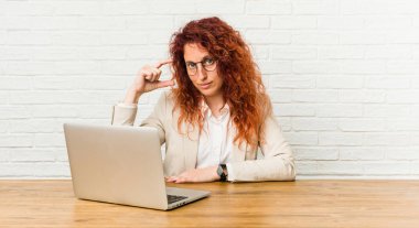 Genç kızıl saçlı kıvırcık kadın dizüstü bilgisayarıyla çalışıyor, işaret parmaklarıyla küçük bir şey tutuyor, gülümsüyor ve kendinden emin..