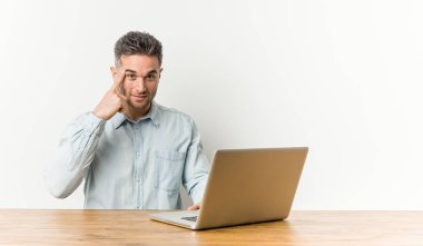 Dizüstü bilgisayarıyla çalışan genç yakışıklı bir adam şakağı parmağıyla gösteriyor, düşünüyor ve bir göreve odaklanıyor..