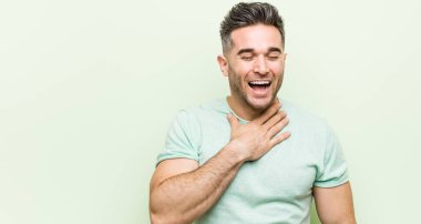Young handsome man against a green background laughs out loudly keeping hand on chest. clipart