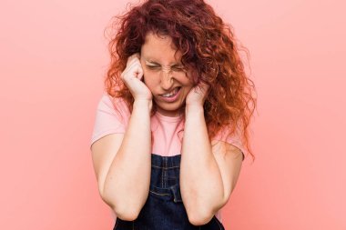 Genç, kızıl saçlı, kot pantolon giyen, kulaklarını elleriyle kapatan bir kadın..