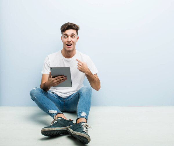 Young man holding a tablet sitting indoor surprised pointing at himself, smiling broadly.