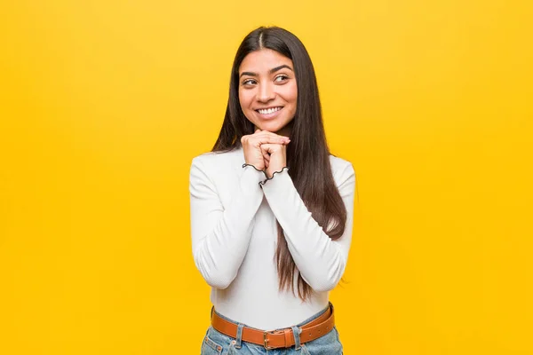 Young pretty arab woman against a yellow background keeps hands under chin, is looking happily aside.