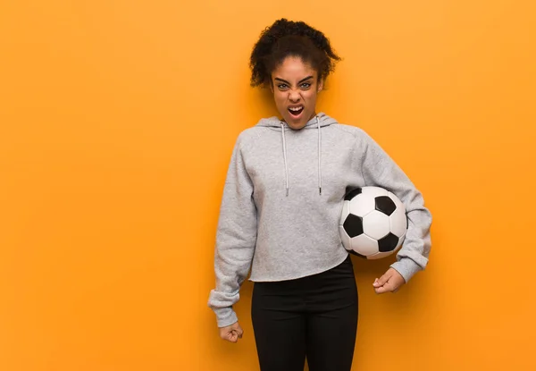Young fitness black woman screaming very angry and aggressive. Holding a soccer ball.