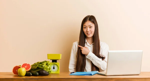 Young nutritionist chinese woman working with her laptop smiling and pointing aside, showing something at blank space.