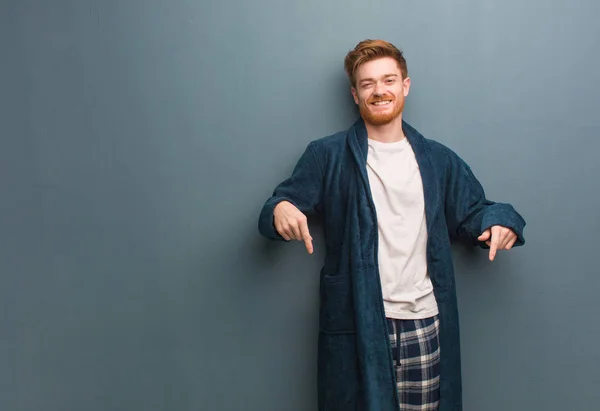 Young redhead man in pajama pointing to the bottom with fingers