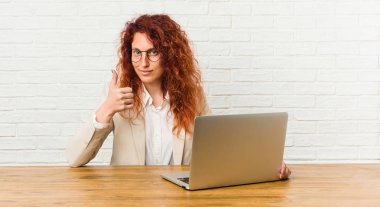 Genç, kızıl saçlı, laptopuyla çalışan, gülümseyen ve baş parmağını kaldıran bir kadın.