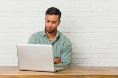 Genç Filipinli adam dizüstü bilgisayarıyla çalışıyor, suratı asık, kolları bağlı..