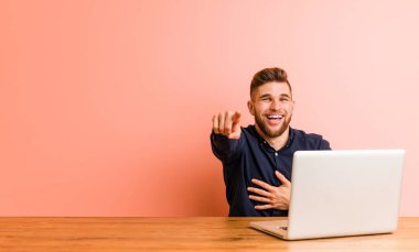 Genç adam dizüstü bilgisayar puanlarıyla çalışıyor, başparmak uzakta, gülüyor ve kaygısız..