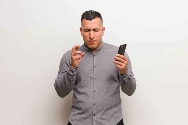 Jovem Latino Segurando Telefone Cruzando Dedos Para Ter Sorte — Fotografia de Stock