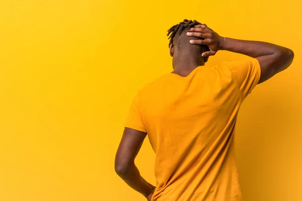 Young black man wearing rastas over yellow background from behind thinking about something.