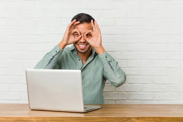 Young filipino man sitting working with his laptop showing okay sign over eyes