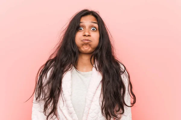 Young indian woman wearing pajama blows cheeks, has tired expression. Facial expression concept.