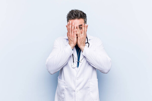 Young handsome doctor man blink through fingers frightened and nervous.