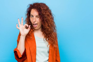 Genç kızıl saçlı zarif bir kadın göz kırpıyor ve elleriyle iyi bir jest yapıyor..