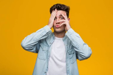 Kot gömlek giyen genç ve yakışıklı bir öğrenci parmaklarının arasından korkmuş ve gergin bir şekilde göz kırpıyor..