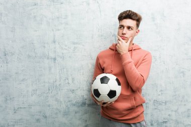 Elinde futbol topu tutan genç bir adam şüpheli ve şüpheci bir ifadeyle yana bakıyor..