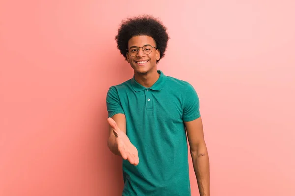 Young african american man over a pink wall reaching out to greet someone