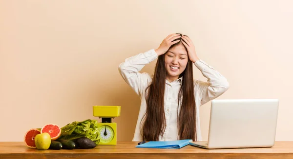 Young nutritionist chinese woman working with her laptop laughs joyfully keeping hands on head. Happiness concept.
