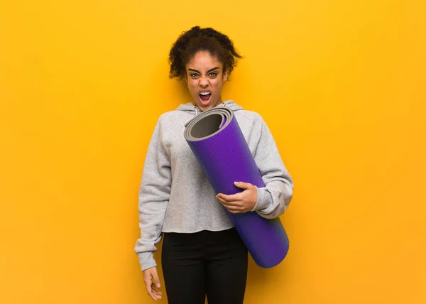 Young fitness black woman screaming very angry and aggressive. Holding a mat.