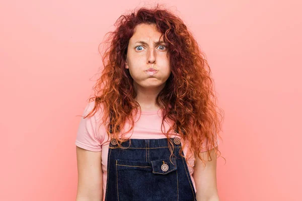 Young pretty ginger redhead woman wearing a jeans dungaree blows cheeks, has tired expression. Facial expression concept.
