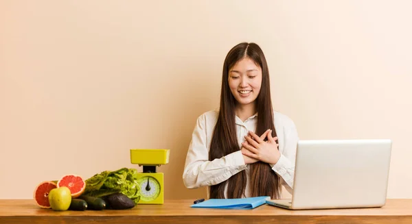 Young nutritionist chinese woman working with her laptop laughing keeping hands on heart, concept of happiness.