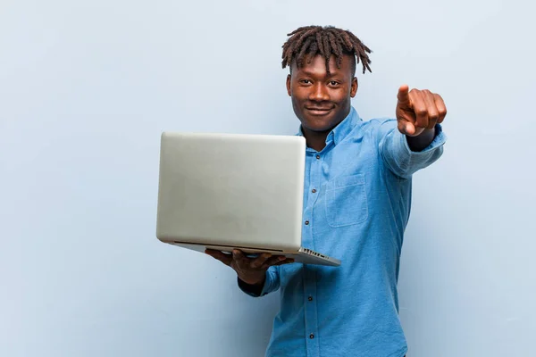 Young rasta black man holding a laptop cheerful smiles pointing to front.