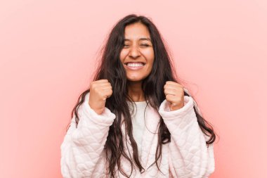 Genç Hintli kadın pijama giyiyor, yumruğunu kaldırıyor, mutlu ve başarılı hissediyor. Zafer konsepti.