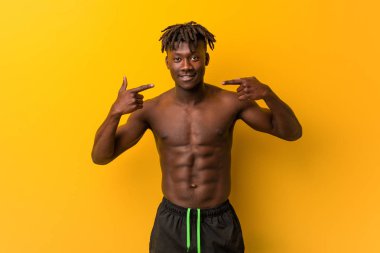 Genç siyah adam, mayo giymiş, gülümseyen, parmaklarını ağzına doğru gösteren..