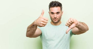 Yeşil arka planda genç yakışıklı bir adam baş parmağını kaldırıyor ve baş parmağını aşağı gösteriyor, zor seçim konsepti.