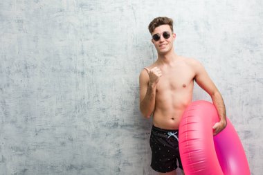 Elinde pembe şişme bir donut tutan, mayo giyen, gülümseyen ve boş bir alanı gösteren genç bir adam..