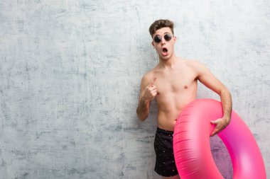 Elinde pembe şişme bir çörek tutan genç adam mayo giyip yan tarafı işaret ediyor.