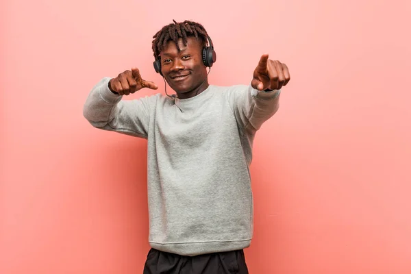 Young rasta black man listening to music with headphones cheerful smiles pointing to front.