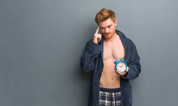 Young redhead man wearing pajama thinking about an idea. He is holding an alarm clock.