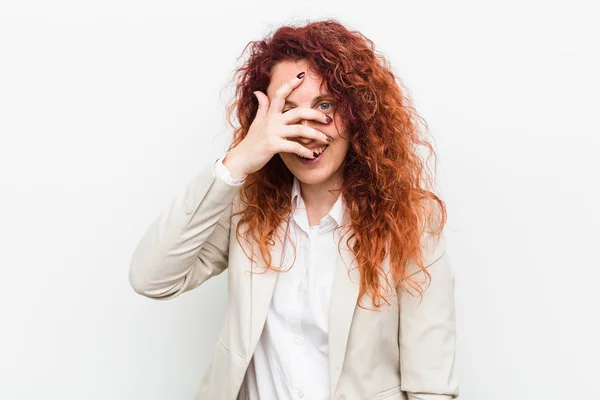 Young natural redhead business woman isolated against white background blink at the camera through fingers, embarrassed covering face.