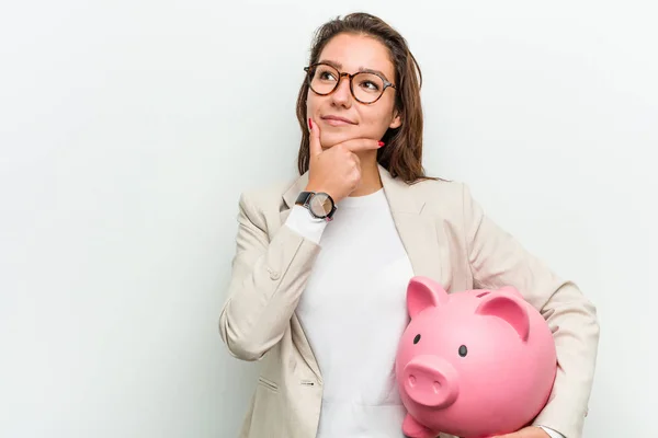 Young European Business Woman Holding Piggy Bank Looking Sideways Doubtful — Stock Photo, Image