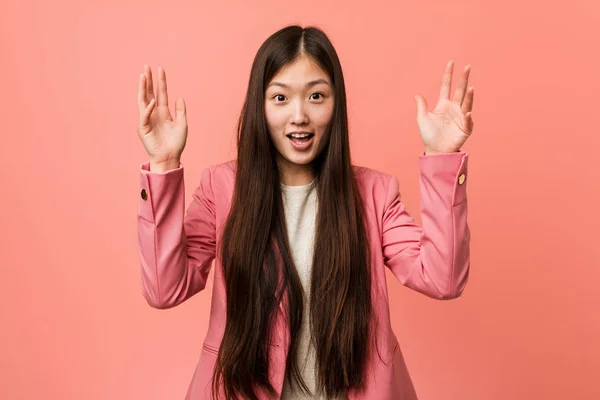 Young business chinese woman wearing pink suit receiving a pleasant surprise, excited and raising hands.