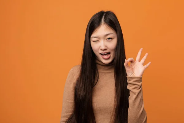 Young pretty chinese woman winks an eye and holds an okay gesture with hand.