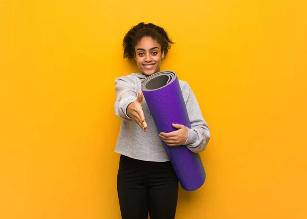 Young fitness black woman reaching out to greet someone. Holding a mat.